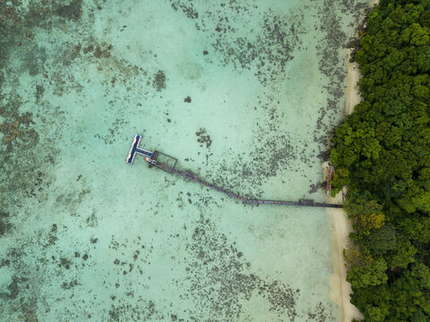 Aerial view of a wooden pier stretching into turquoise waters, contrasting with the lush green shoreline at Turtle Conservation, Karimunjawa, Central Java, Indonesia.