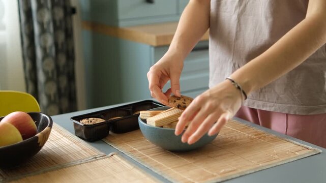 Close up view of female hands arranging a delicious snack, putting chocolate chip cookies and sweet wafers from their packaging into a stylish blue bowl on the kitchen table