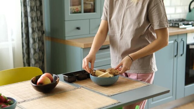 Young woman in pajamas standing at the kitchen table in the morning, taking oatmeal cookies from a package and putting them in a bowl with butter for a delicious and healthy breakfast