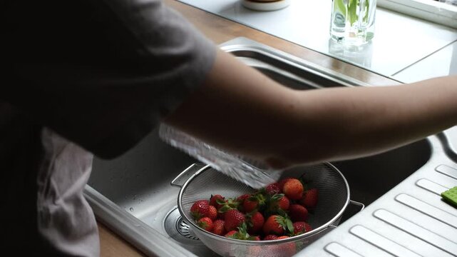 Young woman's hands rinsing fresh ripe red strawberries in a metal colander under running tap water in a modern stainless steel kitchen sink, preparing a healthy fruit snack or dessert