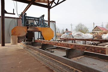 Fototapeta premium Wooden log is cut on machine. Man controls machine for sawing logs. Worker at machine. Production of wooden pallets for heavy cargo transportation. An open-air locksmith workshop.