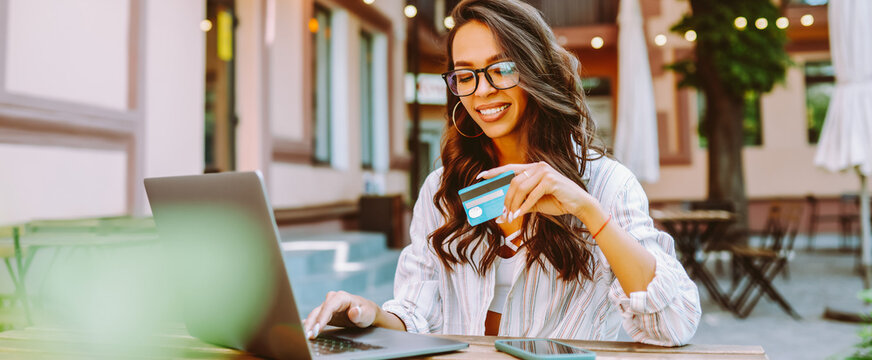 Portrait of woman holding bank card while sitting on cozy terrace in cafe. Young woman wearing glasses and using laptop makes online purchase with debit card. Concept of online shopping.
