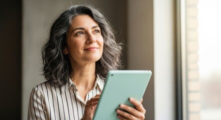 Mature woman with graying hair looks upward thoughtfully while holding a digital tablet near a window