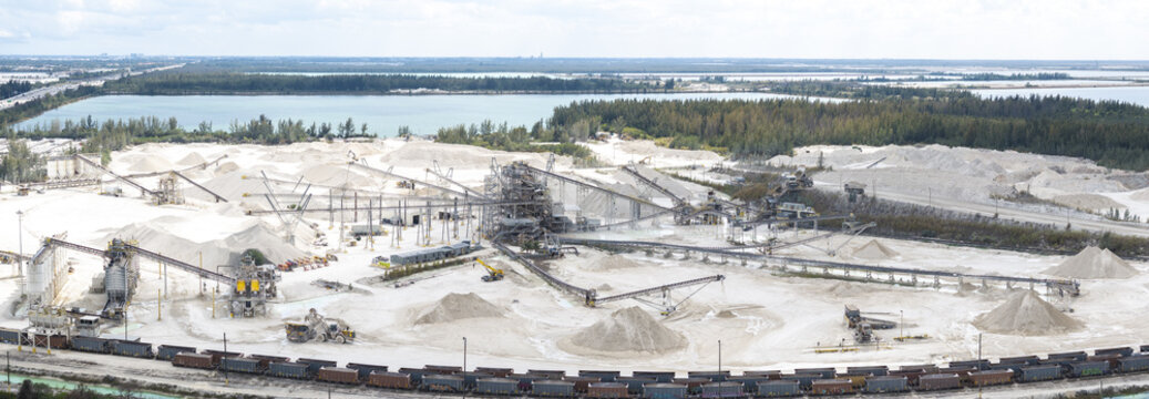 Aerial view of a vast, white limestone quarry contrasting against the blue waters and green vegetation, industrial machinery and trains present, Miami, Florida, United States.