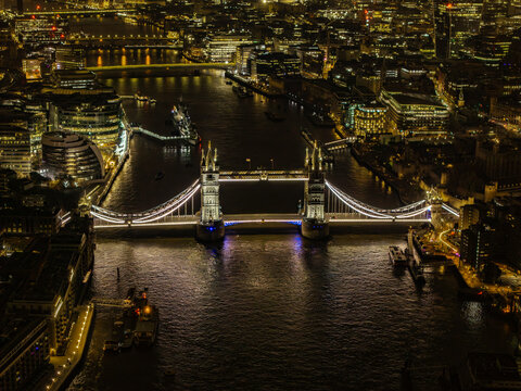 Aerial view of the illuminated Tower Bridge, a jewel amidst the dark Thames, reflecting the city's vibrant nightscape, London, Greater London, United Kingdom.