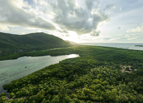 Aerial view of sunlight piercing through clouds over the mangrove forest, casting shadows on the lush greenery meeting the turquoise waters, Karimunjawa, Central Java, Indonesia.