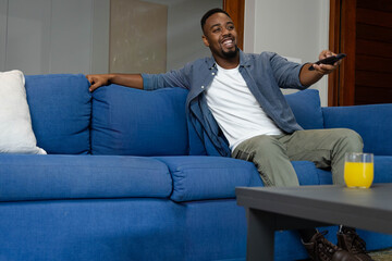 Adult African American man reclining on blue sofa holding remote, orange juice on table, copy space © wavebreak3