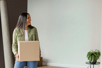 Naklejka premium Mid-adult Indian woman holding cardboard box, wearing green jacket and jeans in minimal room