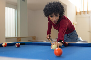 Fototapeta premium Adult African-American woman in jeans leaning over home pool table holding cue lining up cue ball