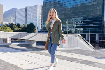 Woman wearing olive parka walking and talking on smartphone gesturing tiled plaza by glass building © wavebreak3