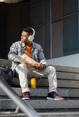 African American man sitting on steps holding bag, listening with headphones, juice beside backpack © wavebreak3