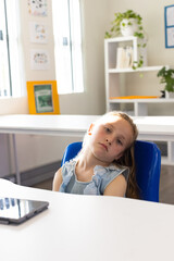 Girl child sitting in blue chair at classroom desk, wearing sleeveless lace top, using tablet