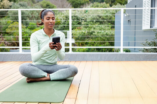 Adult Indian woman sitting crosslegged on green yoga mat on deck in activewear using smartphone