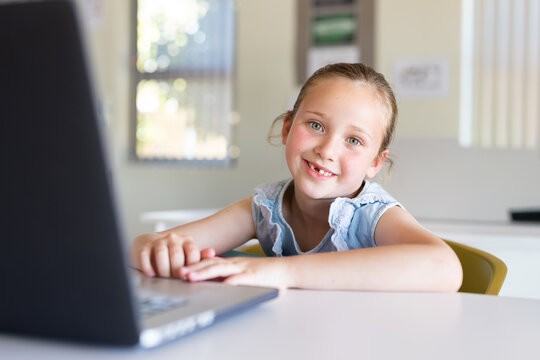Girl sitting at table in classroom working on laptop, smiling with missing tooth, wearing blue top