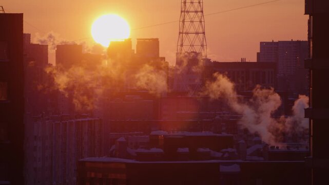 Golden sun rising over a frosty urban cityscape on a cold winter morning, with steam and smoke billowing from building chimneys against a clear sky, highlighting industrial activity
