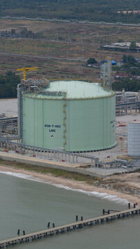Aerial view of a vast LNG storage tank contrasting with the brown landscape and the pier extending into the water, Map Ta Phut, Rayong, Thailand.