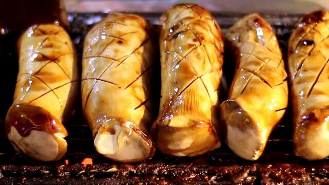 Close up of a vendor brushing BBQ sauce on thick grilled King Oyster mushrooms at a Thai night market. A healthy plant based street food option with a meaty texture and smoky aroma.