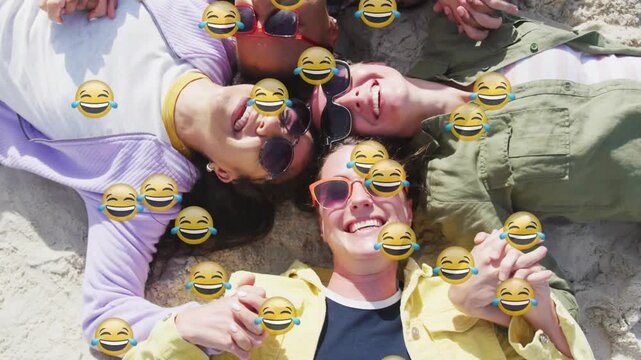 Vertical video: Lying women wearing shades holding hands laughing on sand, emoji overlay appearing