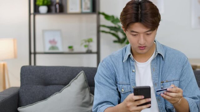 Stressed Young Asian Man Holding Credit Card and Smartphone in Living Room