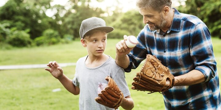 A man teaches a boy baseball in a park. The man, wearing a plaid shirt, shows the boy, in a cap, how to hold a baseball. Both wear gloves. Baseball lesson outdoors. Baseball, sport concept.