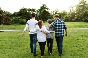 Fototapeta premium A group of four people walking in a park, seen from behind. They are enjoying a sunny day, surrounded by greenery and trees, embracing each other. Happy family group photography in park.