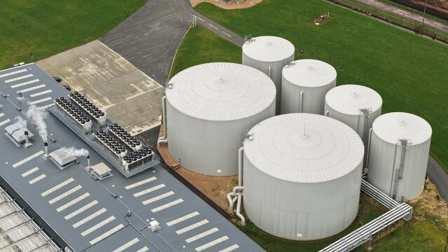 Aerial view of cylindrical tanks gleam under the soft light alongside a building emitting plumes of white smoke, Fenland Glasshouse Energy Centre, Chittering, Cambridge, United Kingdom.