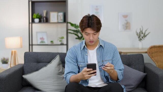 Stressed Young Asian Man Holding Credit Card and Smartphone in Living Room