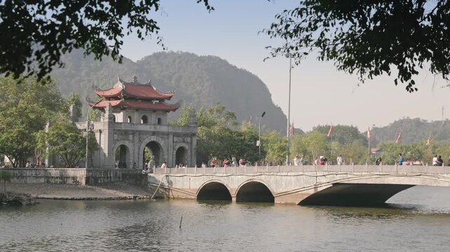 People stroll across a bridge to visit the  Hoa Lu Ancient Capital, Ninh Binh, Vietnam. The bridge spans a lake, offering scenic views of the surrounding mountains and landscape.