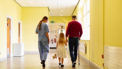 A nurse, a man with a cane, and a child walk down a bright hallway. The nurse assists, the man uses a cane, and the child holds a teddy bear. Healthcare support concept, hospital setting.