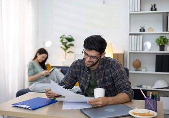 Obraz premium A young man smiles as he reviews documents on his laptop, while his partner relaxes behind him. The atmosphere is warm and inviting in this home office, reflecting the concept of working from home.