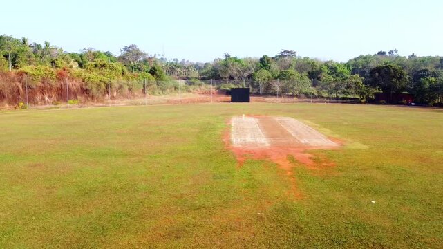 Aerial wide shot of a rural cricket field with a central pitch, surrounded by lush tropical greenery and a practice net facility under a clear sky.