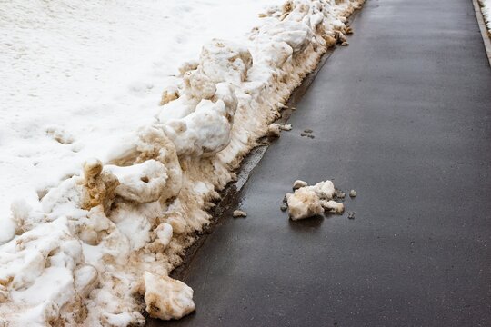 Grunge melting snow piled against a dark, wet asphalt path. Muddy slush and puddles signal the arrival of an early spring thaw, ending winter's grip
