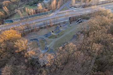 Fototapeta premium Aerial view of a bike pump track and forest recreation area next to an urban road at sunset.