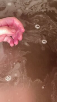Hands holding purple bomb over water, closeup of fizzing bomb dissolving into tub, vibrant purple pigment releasing into water,