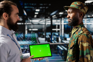 Fototapeta premium Male army personnel and technician using chroma key display on tablet, troubleshooting AI systems for a national safety military operations. Officer and server farm expert on industrial platform.