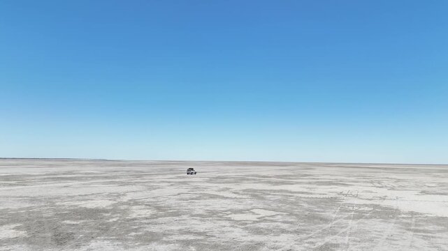 Aerial drone footage captures a lone car driving across the vast, white expanse of the Makgadikgadi Pans in Botswana, heading toward the iconic Kubu Island.