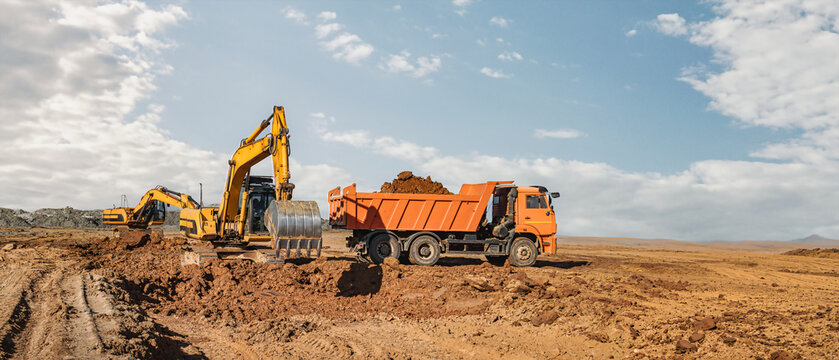 Heavy machinery is moving dirt at a construction site. A dump truck is loading soil while an excavator works nearby. The sun shines in a clear sky