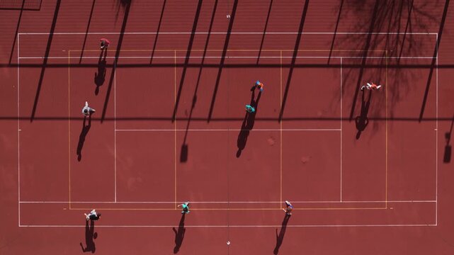 Aerial view of a red tennis court with players casting long shadows, creating a dynamic pattern of light and shade on the vibrant surface, Bratislava, Slovakia.