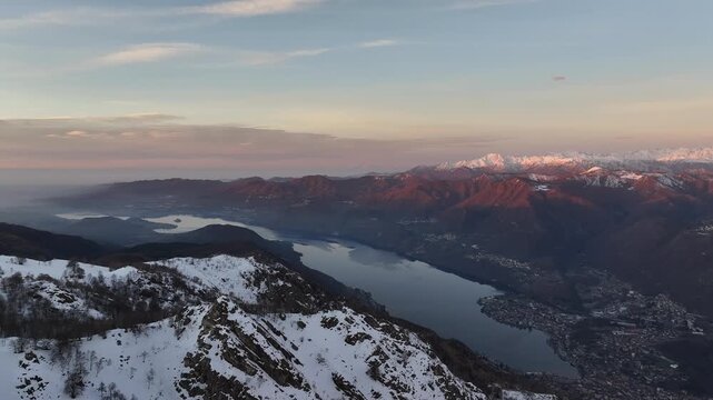 Aerial view of snow-covered Mottarone mountains meeting the tranquil lake waters, painted with the warm hues of the setting sun, Mottarone, Piedmont, Italy.