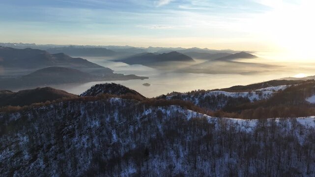 Aerial view of snowy Mottarone mountain ranges and a distant lake, the landscape bathed in the soft glow of the morning sun, Mottarone, Piedmont, Italy.