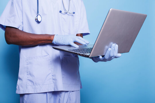 Man working as nurse using wireless computer while wearing stethoscope, gloves for protection and scrubs. African american individual standing and utilizing a digital laptop.