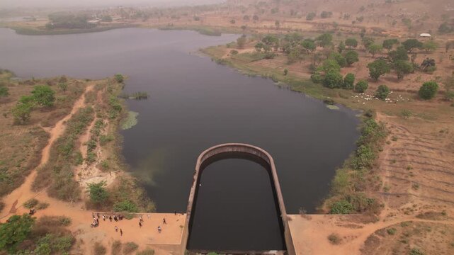 Aerial view of a concrete dam holding back dark water, contrasted against the dry, dusty landscape and sparse vegetation, Suleja, Niger, Nigeria.