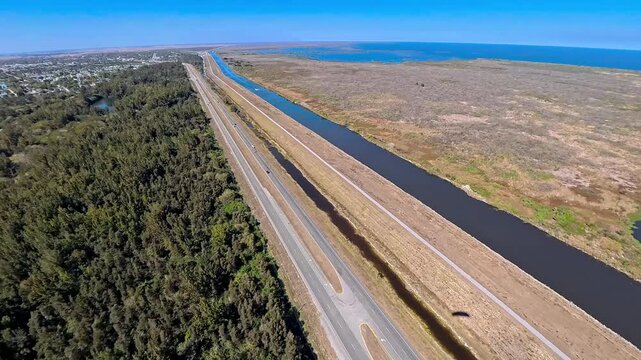 Aerial view of a straight highway and canal cutting through lush green trees and marshland under a clear blue sky, Lake Harbor, Florida, United States.