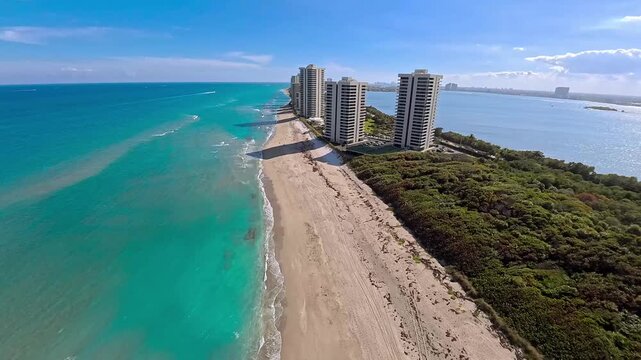 Aerial view of beachfront condos casting long shadows on the golden sand, contrasting with the turquoise sea and verdant vegetation, Riviera Beach, Florida, United States.