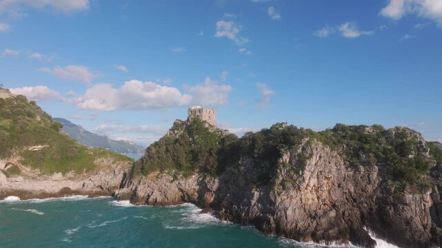 Aerial view of Torre Capo di Conca perched atop a rocky outcrop with lush greenery against the blue sky and sea, Conca dei Marini, Campania, Italy.