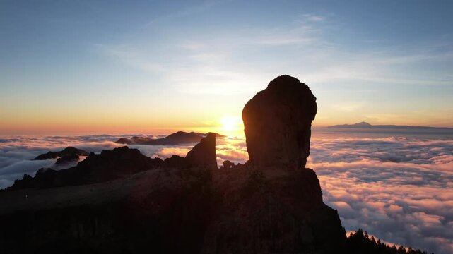 Aerial view of Roque Nublo peak silhouetted against a vibrant sunset, with clouds creating a sea of pink and white, Tejeda, Canary Islands, Spain.