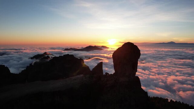 Aerial view of Roque Nublo's dramatic silhouette against the vibrant sunset, a sea of clouds, and distant mountains, Tejeda, Canary Islands, Spain.