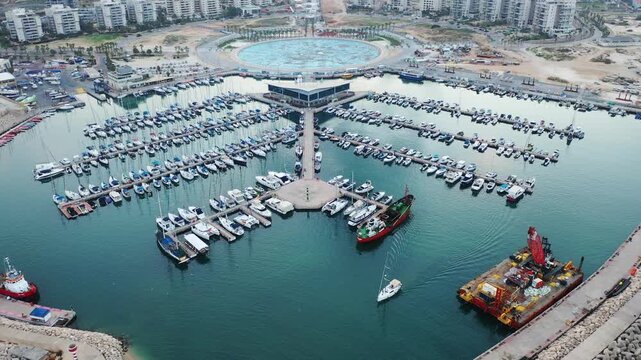 Aerial view of the Ashdod Marina filled with boats, surrounded by modern buildings and a circular structure, creating a vibrant contrast of urban and maritime scenery, Ashdod, South District, Israel.