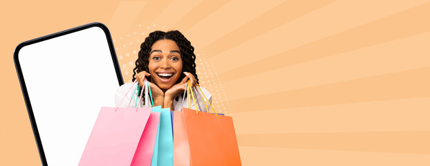 A woman smiles widely while holding colorful shopping bags. She stands in front of a large phone with a blank screen. The activity represents shopping and modern digital interaction. © Prostock-studio