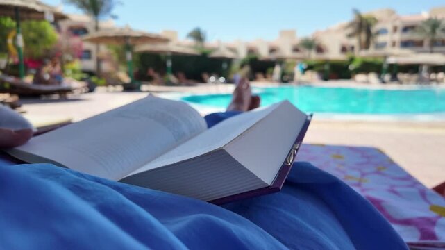 Girl lies on a lounger with an open book by the pool in luxury resort or hotel on a peaceful, sunny day. Slow motion view and copy space for text.